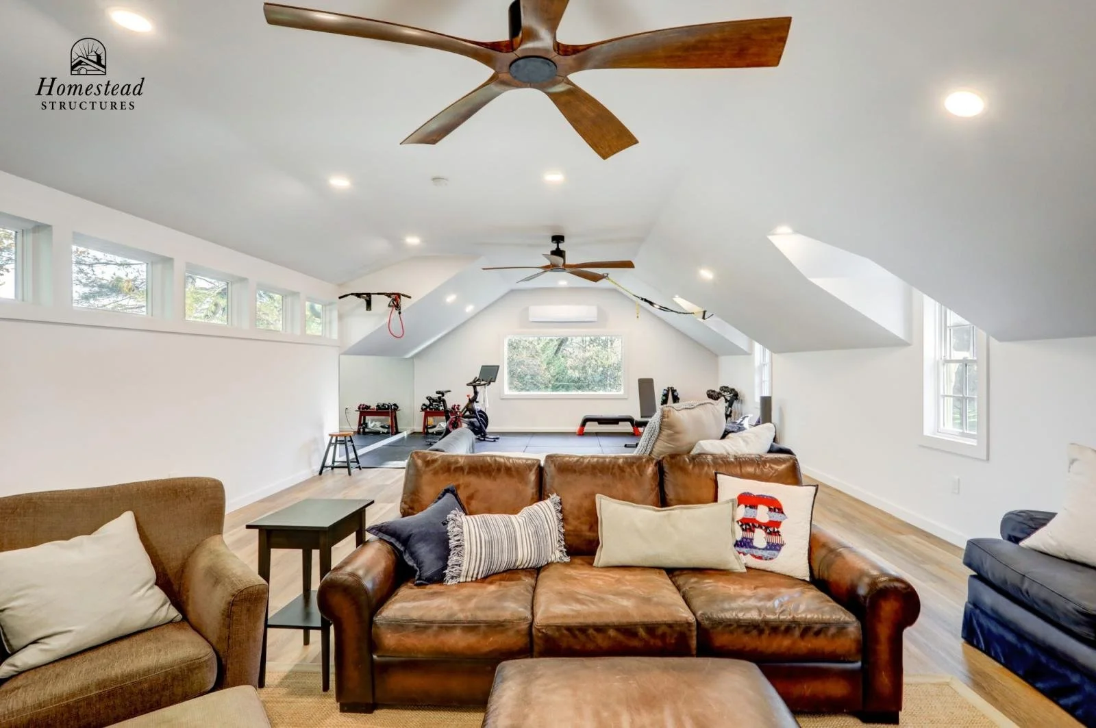Interior of a spacious attic living room with white walls and ceiling. It features a brown leather sofa decorated with throw pillows, a beige armchair, and a small side table. The ceiling has large central and smaller side windows, with ceiling fans 