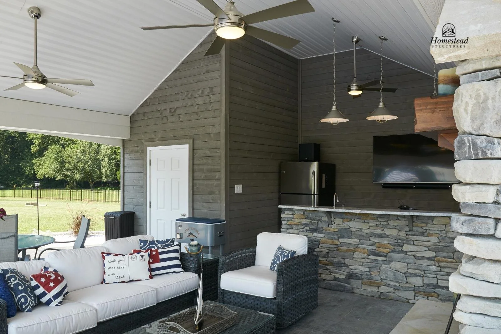 Covered outdoor patio with ceiling fans, a sofa with patriotic pillows, a stone bar counter, and a nearby backyard with trees and a fence.