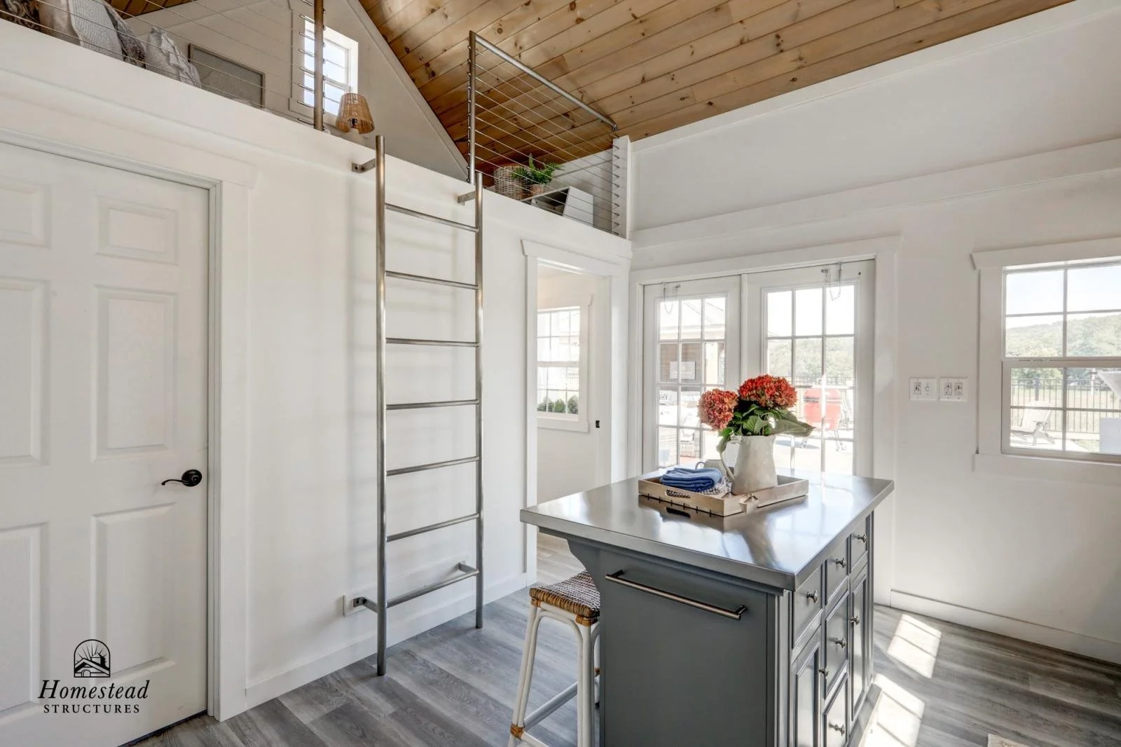 Modern interior of a kitchen with white walls, a small island with a vase of pink flowers on top, large windows letting in natural light, a door, and a metal ladder leading to a loft area with a wooden ceiling and railing.