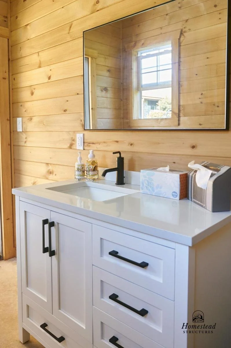 A bathroom vanity with a black faucet, white countertop, and storage drawers and cabinets, set against wood-paneled walls with a large mirror and a window reflected in the mirror.