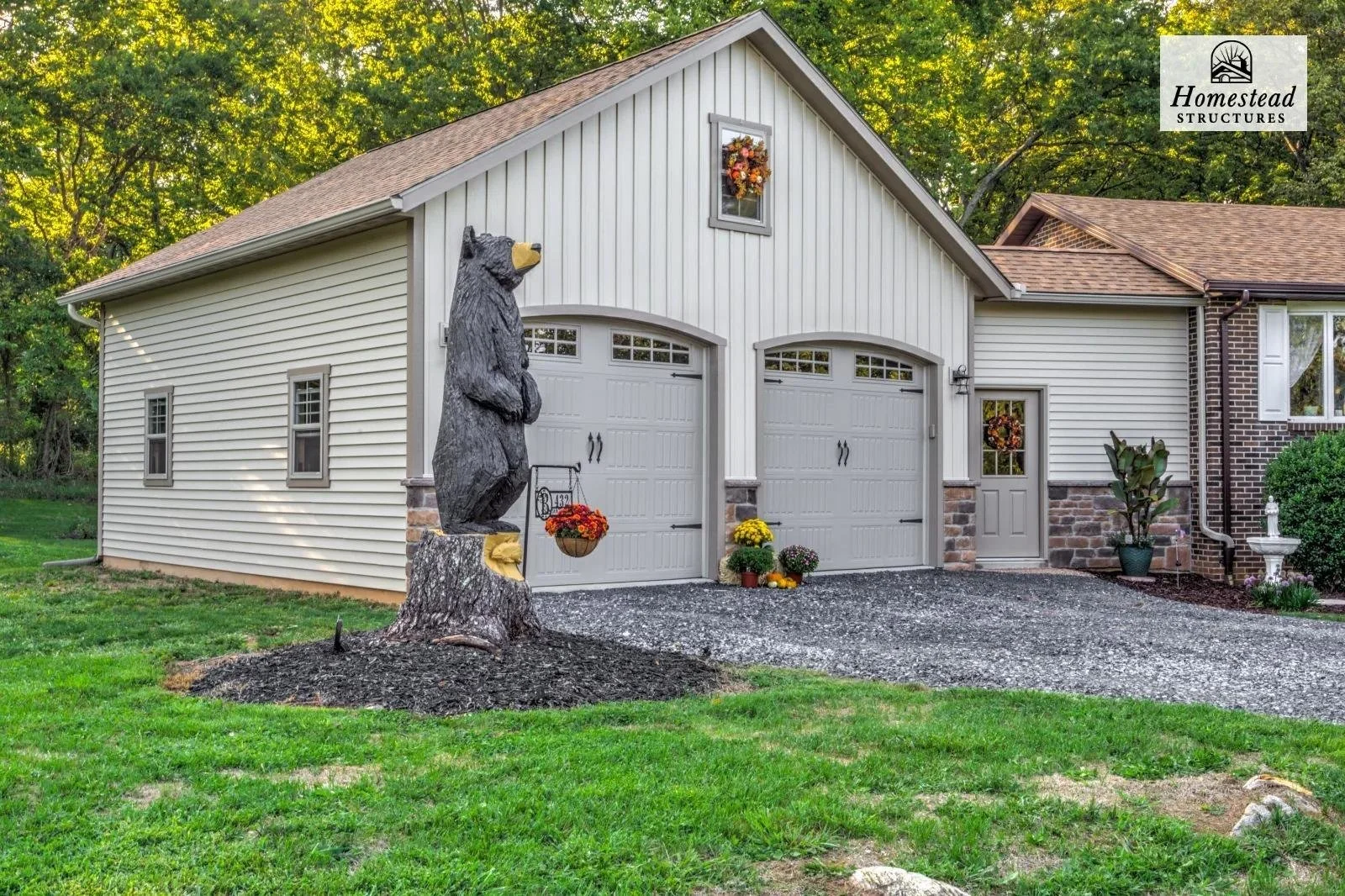 A house with white siding and a stone foundation, decorated for fall with pumpkins and flowers, has a carved bear statue holding a basket hanging from a tree trunk in the front yard. There is a two-car garage, a small side door, and a birdbath. The s