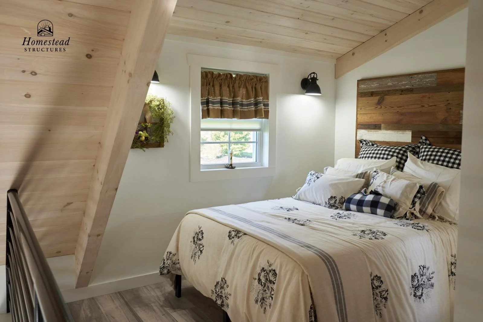 Bedroom featuring a made bed with white and black floral bedspread, plaid pillows, and a wooden headboard. The room has white walls, a window with brown and black valance, black wall sconce, and a wall-mounted plant with flowers.