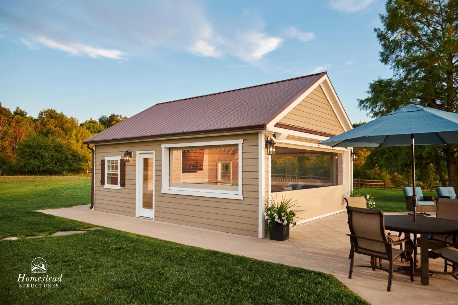 A small, beige house with white trim and a brown metal roof, surrounded by green lawn and trees, with outdoor furniture and an umbrella in the yard.