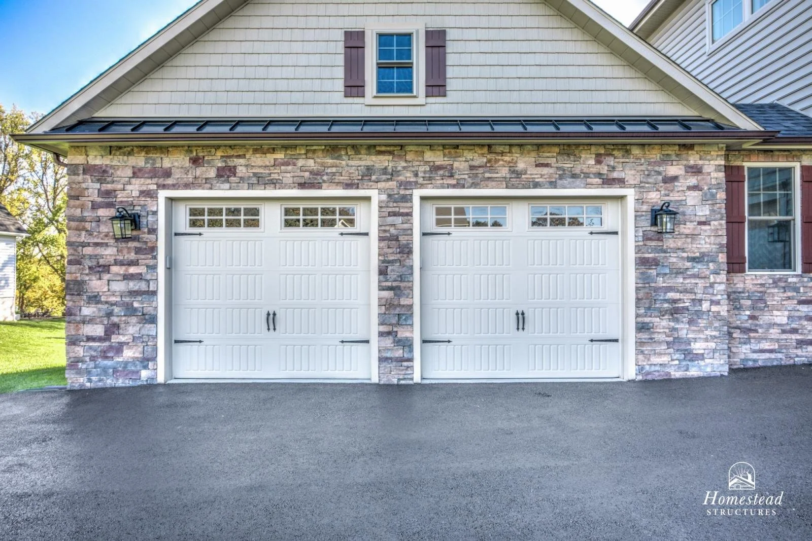 Two white garage doors with small windows at the top, set in a brick building facade with stone accents, flanked by black outdoor wall lanterns, with a black asphalt driveway in front.