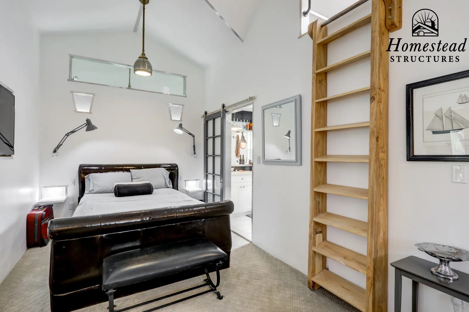 A bedroom with white walls, a dark wood bed with gray and white bedding, two wall-mounted adjustable lamps, a sliding barn door leading to a bathroom, a tall wooden shelf, a small black table with a decorative silver bowl, and small rectangular wall-