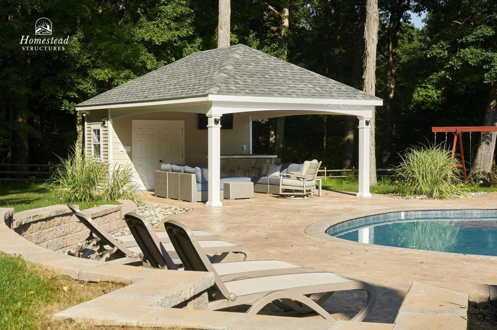 A backyard scene with a swimming pool surrounded by lounge chairs, a gabled pavilion with outdoor seating, and mature trees in the background.