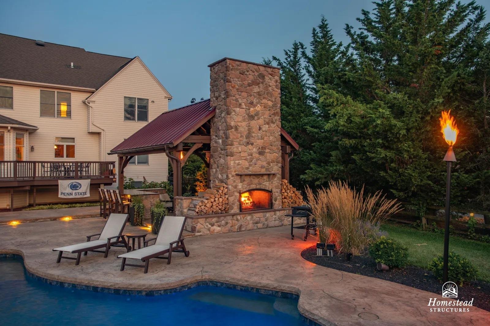 Backyard with outdoor stone fireplace, two lounge chairs near a swimming pool, a torch, and a house in the background.