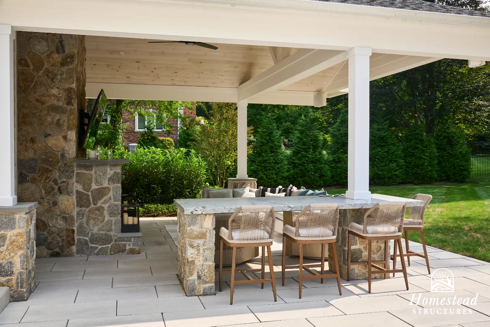 Outdoor patio with stone and concrete bar, four chairs, and surrounding lush green bushes and trees.