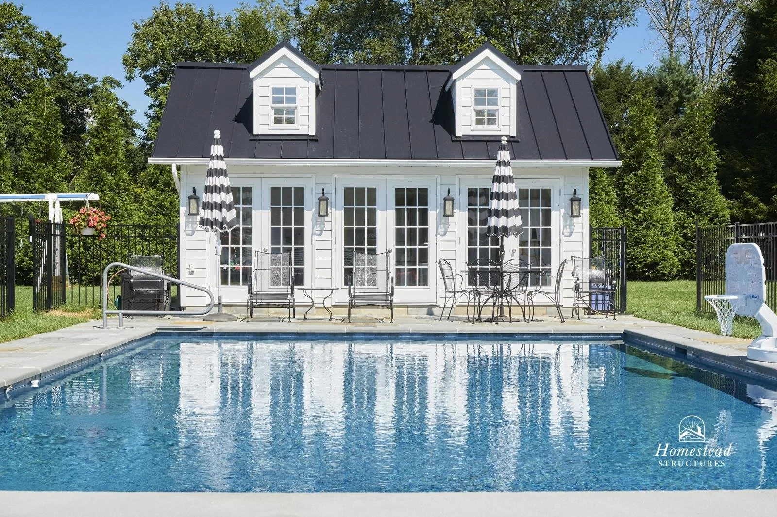 A backyard with a swimming pool in front of a white house with black roof and two dormer windows, surrounded by greenery, with patio furniture and umbrellas.