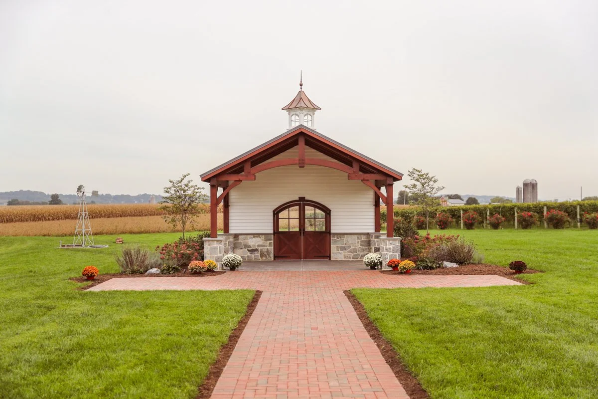 A small church with white walls, a red roof, and a stone foundation, set in a well-maintained grassy area with potted flowers, a brick walkway, and a rural landscape in the background.