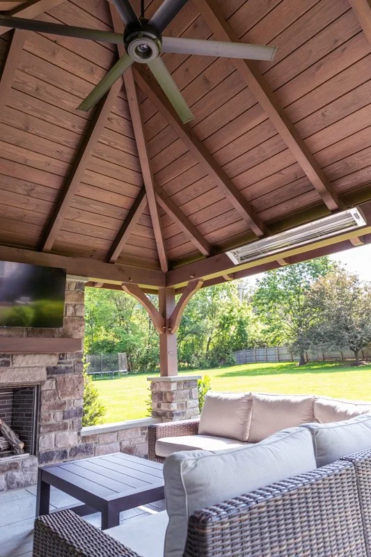 Under a covered patio with a wooden ceiling and a ceiling fan, there is outdoor furniture including a cushioned sofa, a wicker chair, and a small table. The patio overlooks a grassy backyard with trees and a wooden fence.