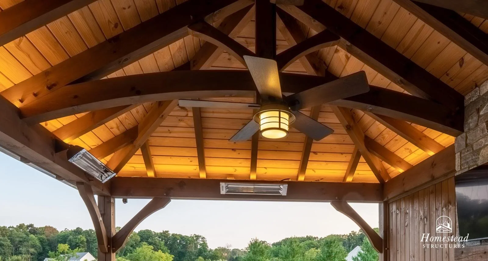 Wooden gazebo ceiling with a modern ceiling fan and built-in lighting, overlooking a green landscape.