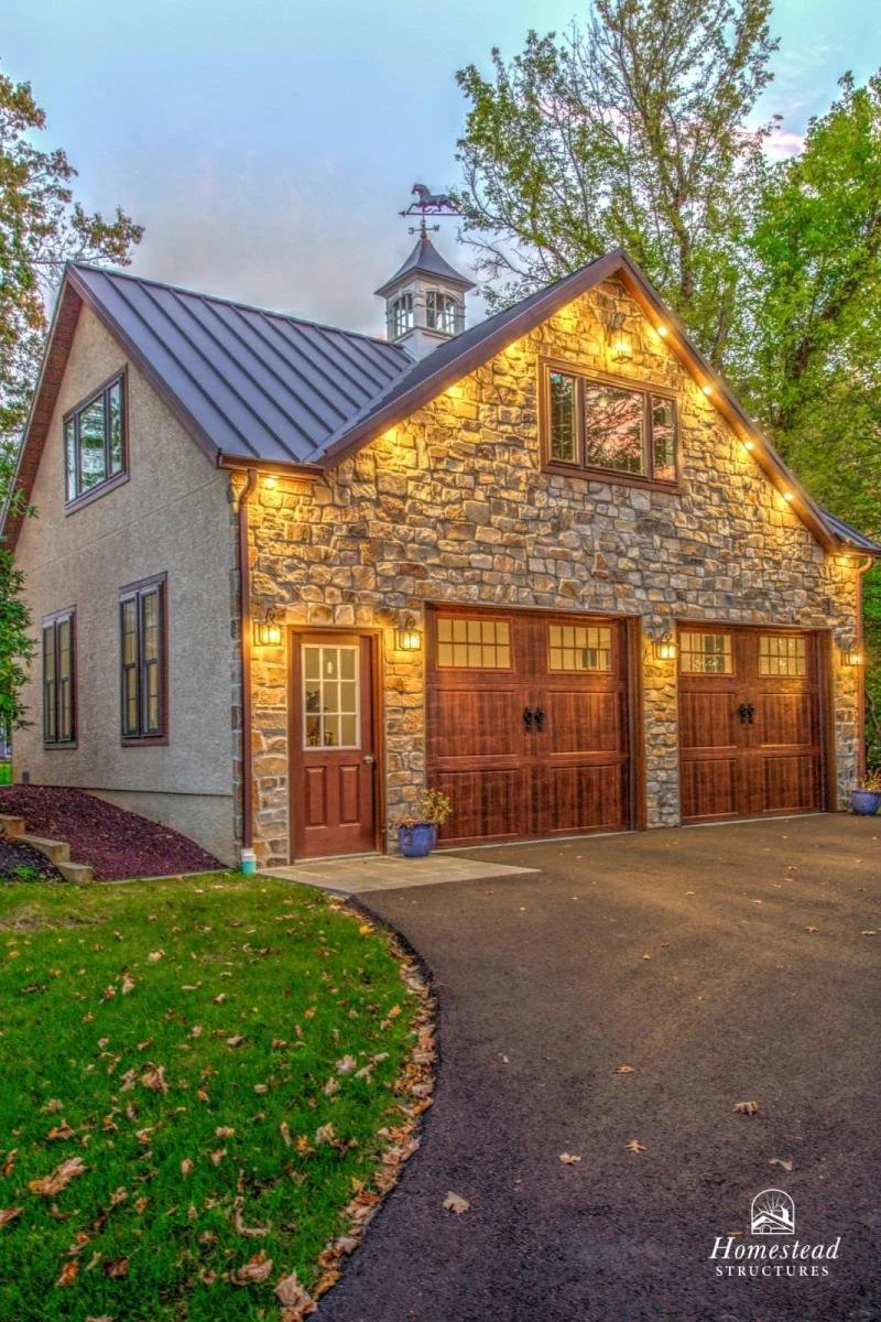 A two-story house with stone and stucco exterior walls, illuminated with outdoor lights, featuring a two-car garage with wooden doors, a small front door, and a decorative weather vane on the roof. Surrounded by trees and a well-maintained lawn.