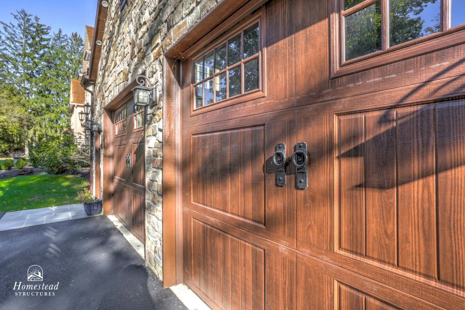 Close-up of a wooden garage door with black handles, stone wall, and outdoor lights on the wall. Green grass and trees in the background.