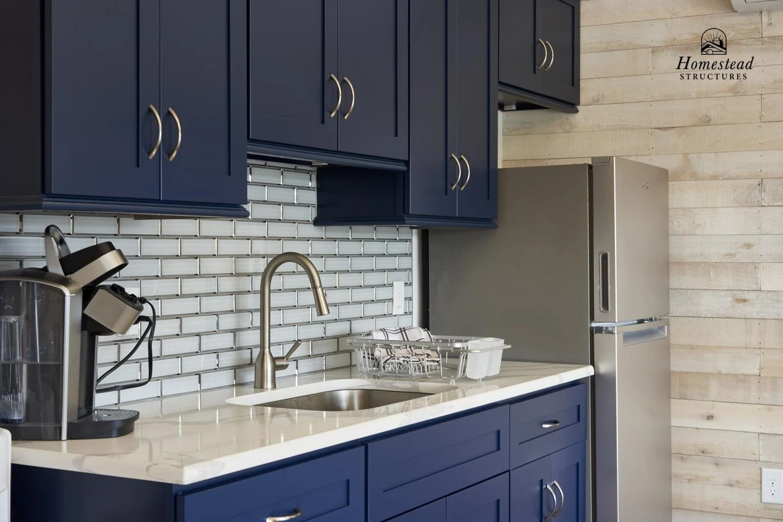 A modern kitchen with blue cabinets, a white countertop, a stainless steel refrigerator, and a white subway tile backsplash. There is a stainless steel faucet above the sink and a black coffee maker on the counter.