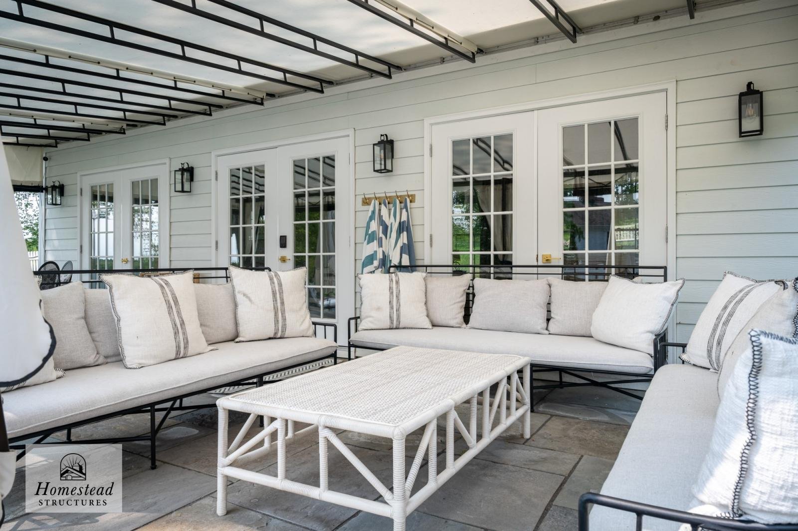 Outdoor patio with beige cushioned seating, black metal frame furniture, and a white woven table in front. White house exterior with three-pane French doors, matching wall lanterns, and a striped towel hanging on a hook.