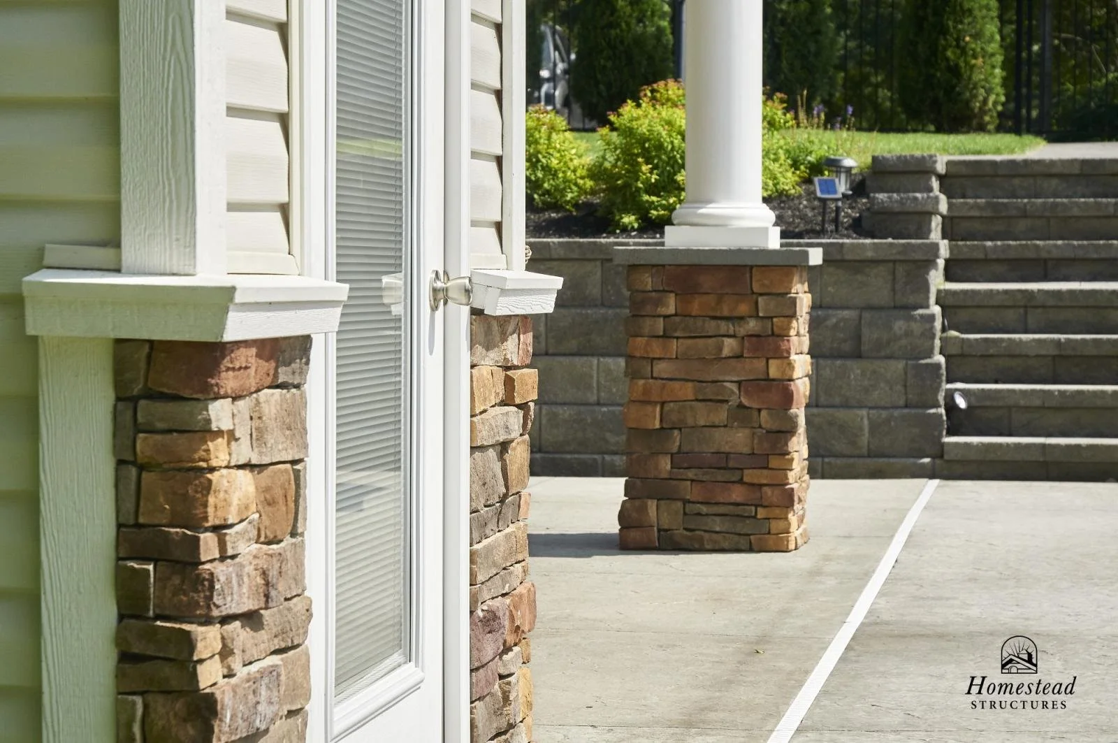 Close-up of a house exterior featuring stone and siding columns, a white door with a handle, brick pillars, and stairs with landscaping in the background.