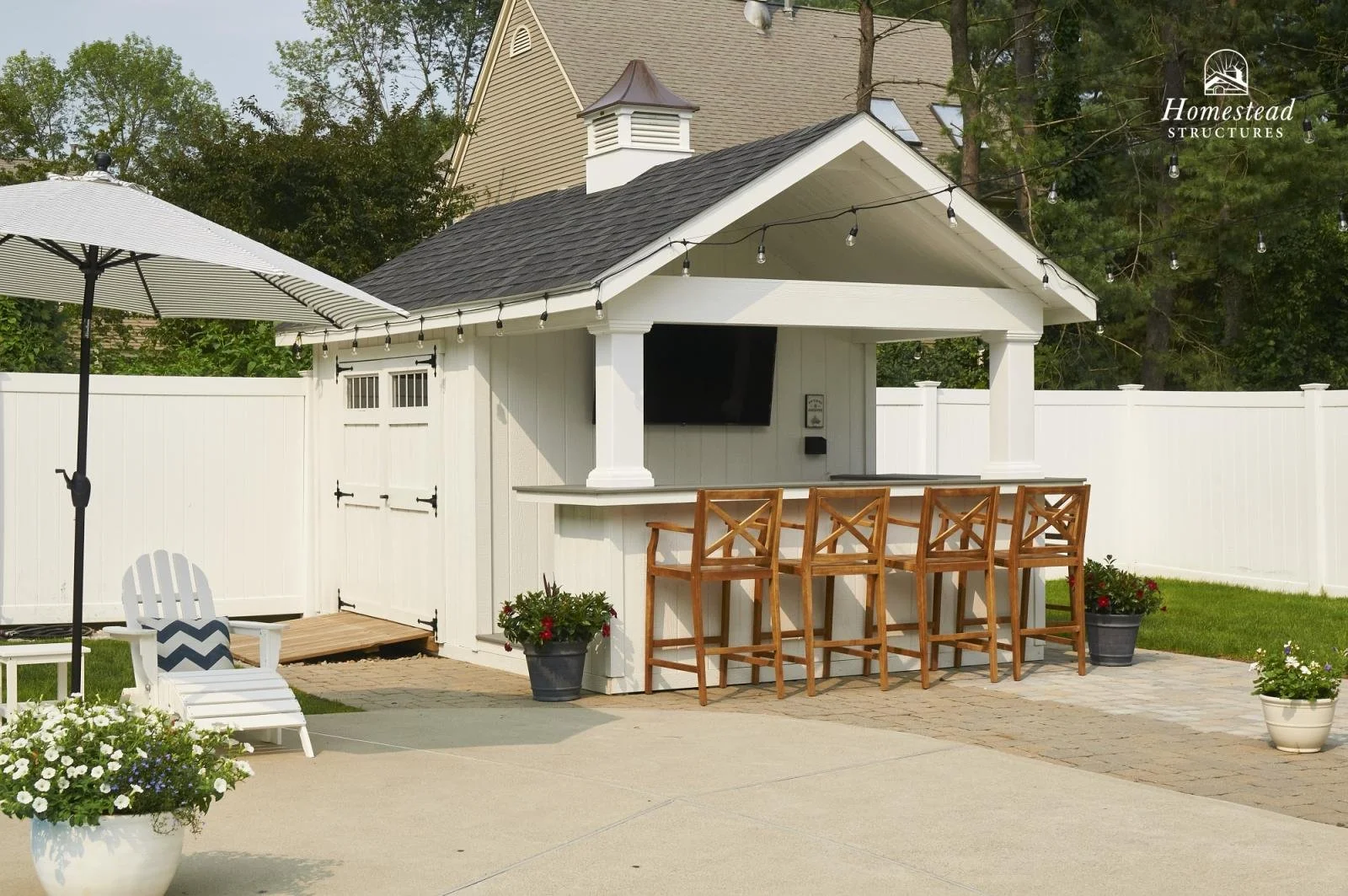 An outdoor backyard bar area with white fencing, a TV, and wooden bar stools. There are potted plants, an umbrella, and string lights hanging above.