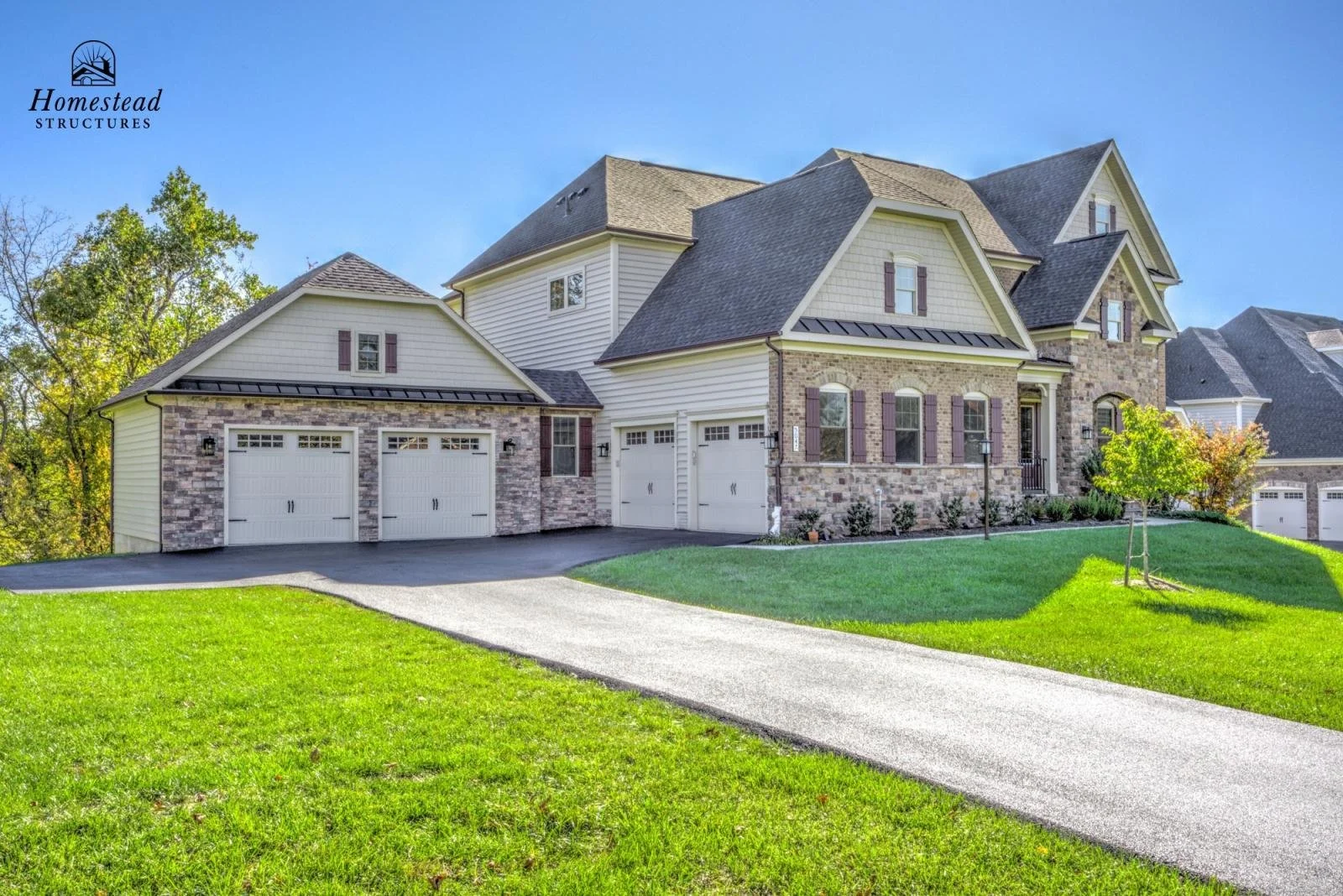 A large, multi-story house with a brick and beige siding exterior, multiple gabled roofs, and a three-car garage with white doors. The house is set on a green lawn with a curved driveway and small trees, under a clear blue sky.