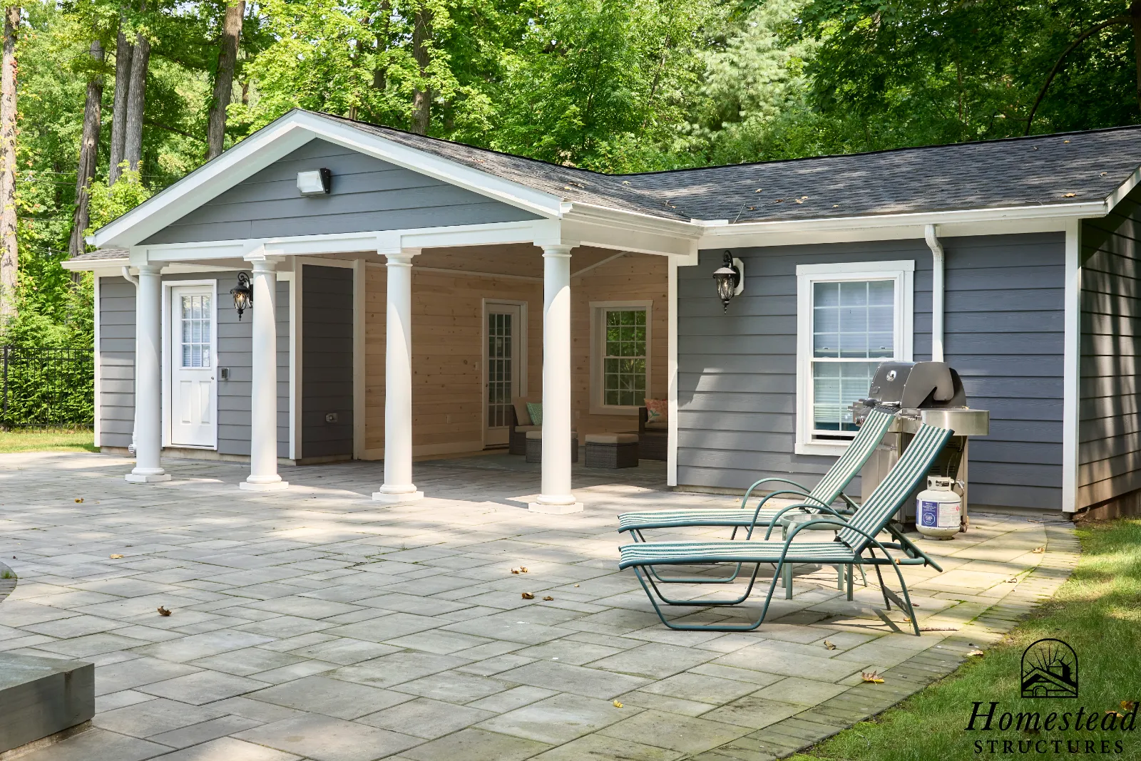 A backyard patio area with a gray house featuring white trim, a covered porch with wooden ceiling, outdoor seating, a gas grill, and green trees in the background.