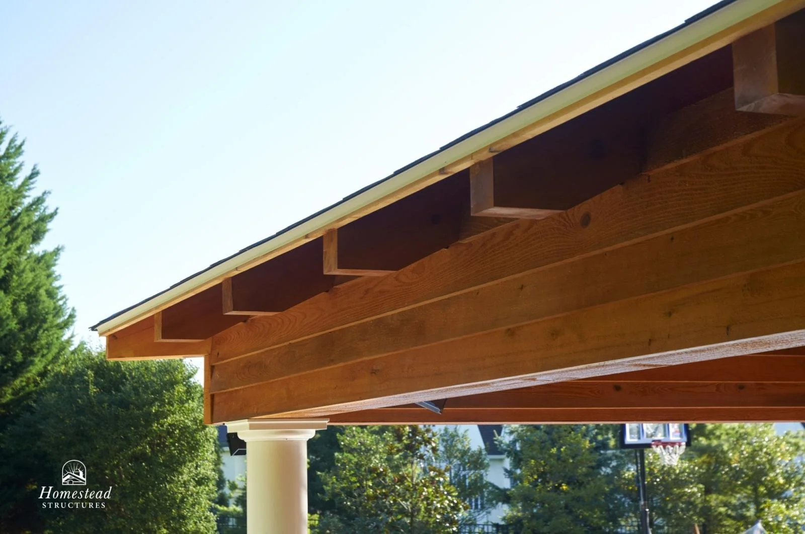 Close-up of a wooden outdoor structure supported by white columns, with a roof under construction, in a backyard with trees and a basketball hoop in the background.