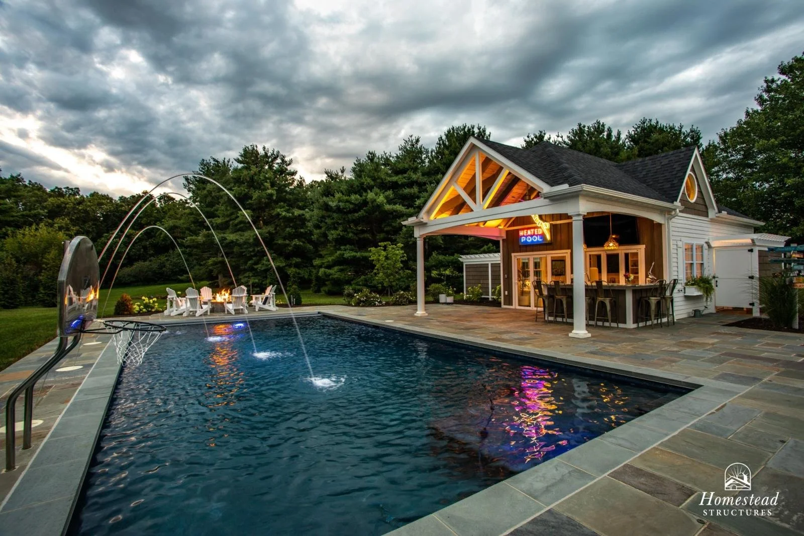 A backyard with a swimming pool, a poolside bar, and a pool house. The pool has water fountains and a basketball hoop. The pool house has a neon sign that says 'Heated Pool' and is surrounded by trees and cloudy sky.