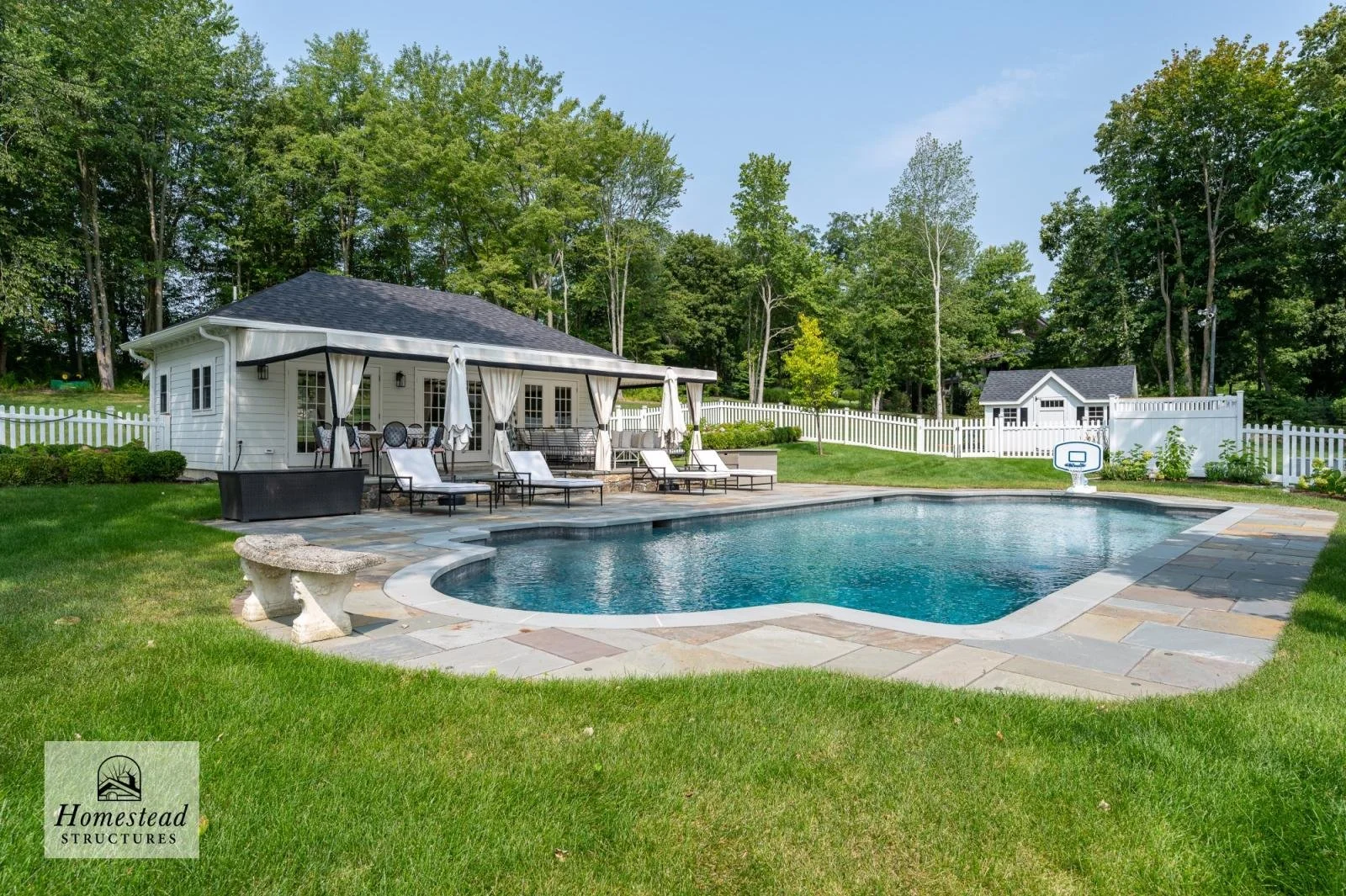 A backyard with a swimming pool, a white pool house with a covered patio, outdoor furniture, decorative curtains, a small soccer goal, a white picket fence, a shed, and surrounding greenery with tall trees.