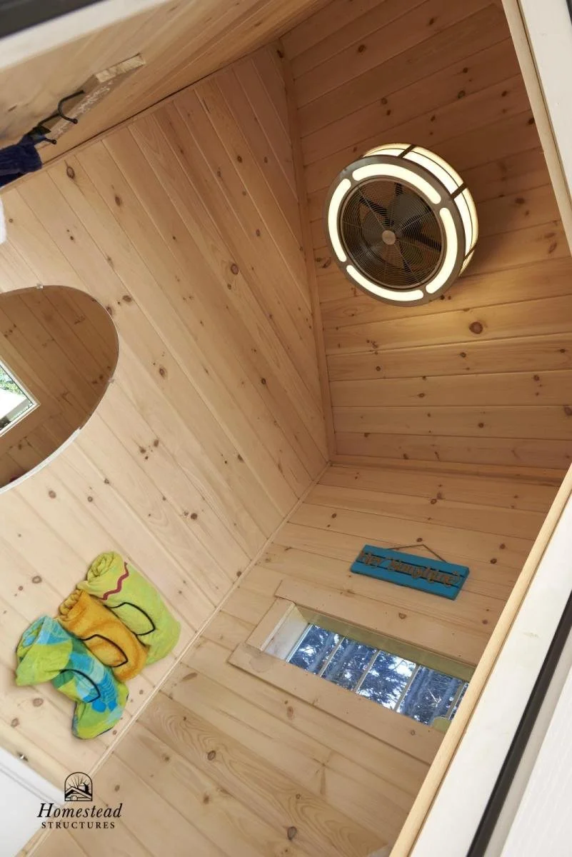 Wood-paneled ceiling with a circular ceiling fan and a small window, colorful towels hanging on the wall, and a blue decorative sign.