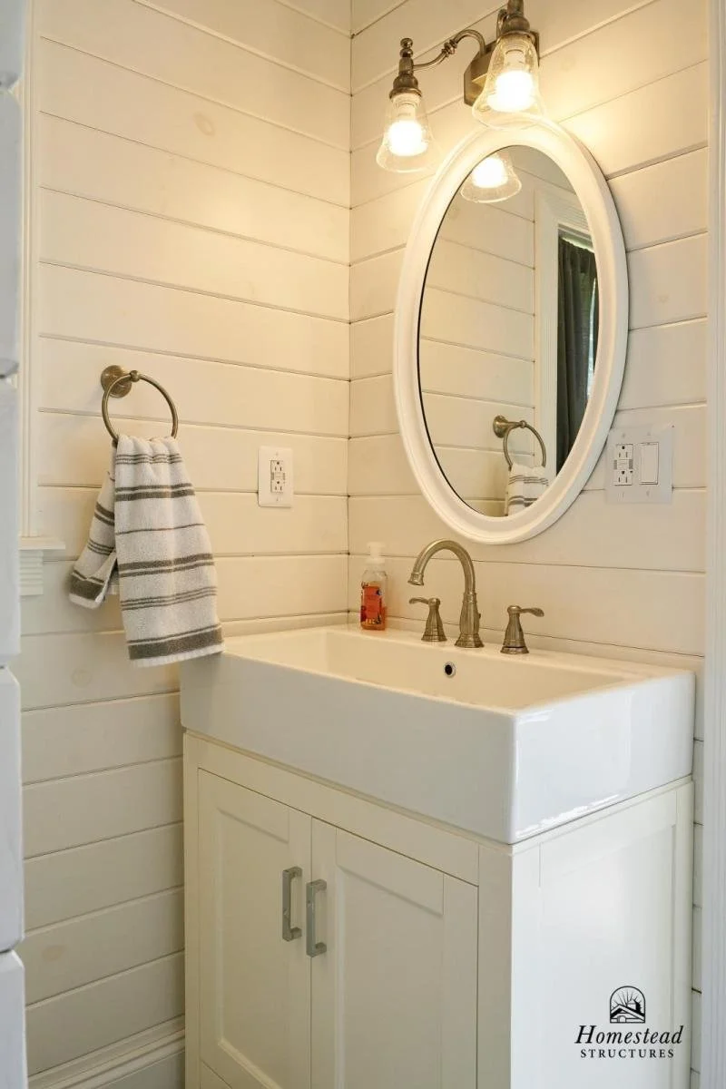 A rustic bathroom with white shiplap walls, a white vanity with a rectangle sink, a silver faucet, and a round mirror. There is a light fixture with three exposed bulbs above the mirror, and a towel ring with a striped towel on the wall. A soap dispe