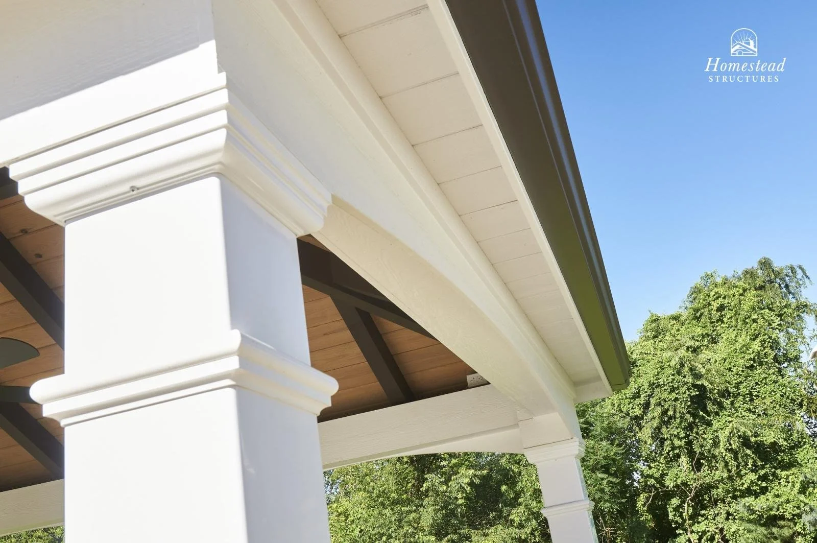 Close-up of the white porch columns and painted trim of a house with a view of green trees and a clear blue sky in the background.