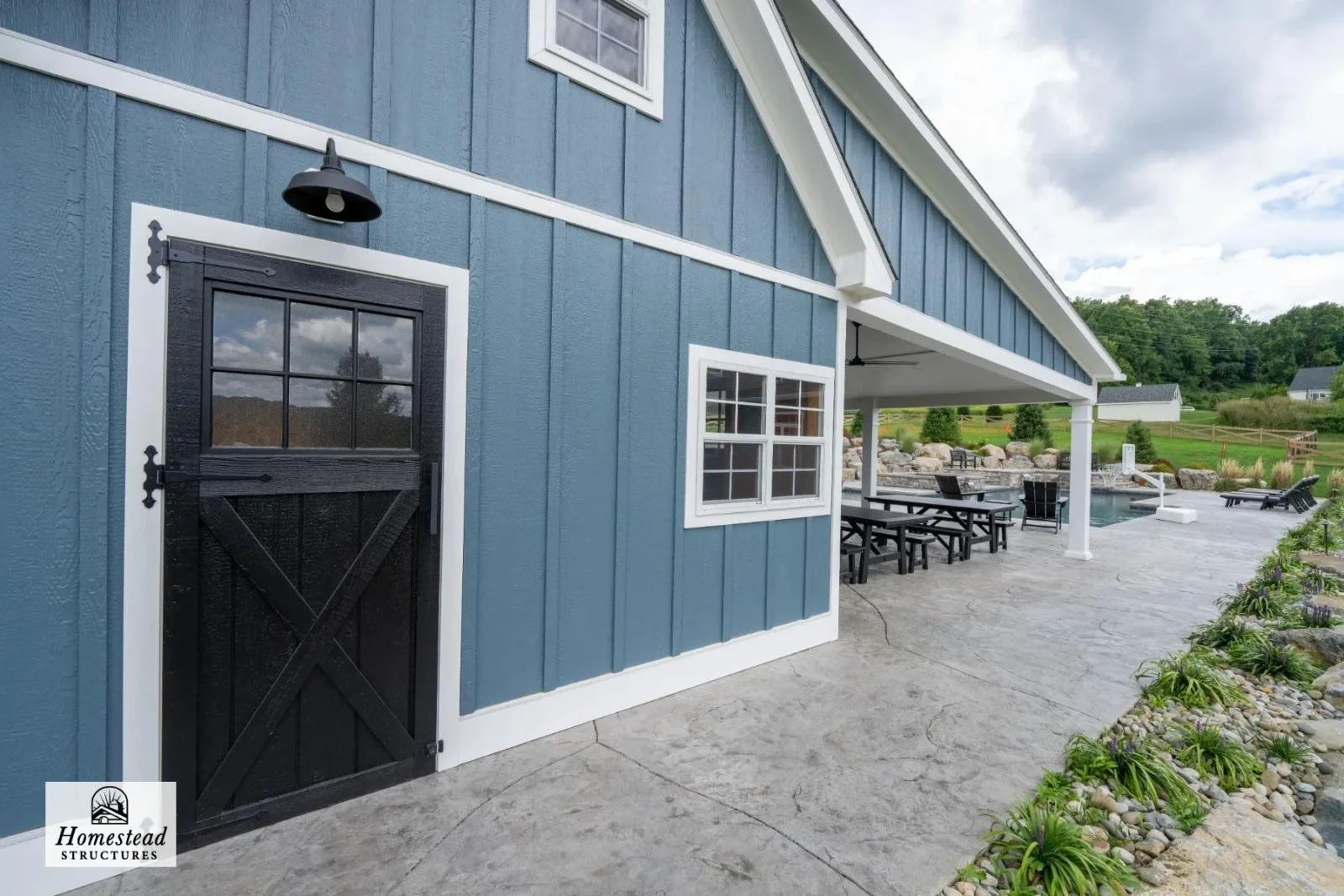 Exterior view of a blue house with a black barn-style door and white trim, showing a covered patio area with outdoor furniture, overlooking a landscaped yard with a pond and greenery.
