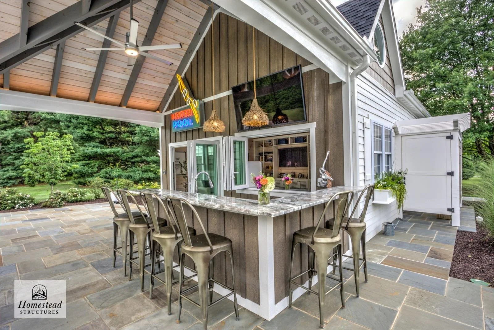 Outdoor patio area with a bar, metal chairs, a TV, a neon sign, and greenery in the background.
