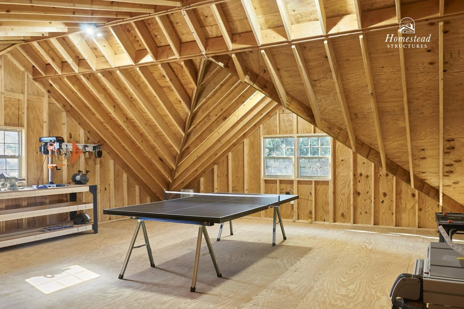 An unfinished attic with wooden walls and ceiling, a black ping pong table, a workbench with tools, and a window letting in natural light.