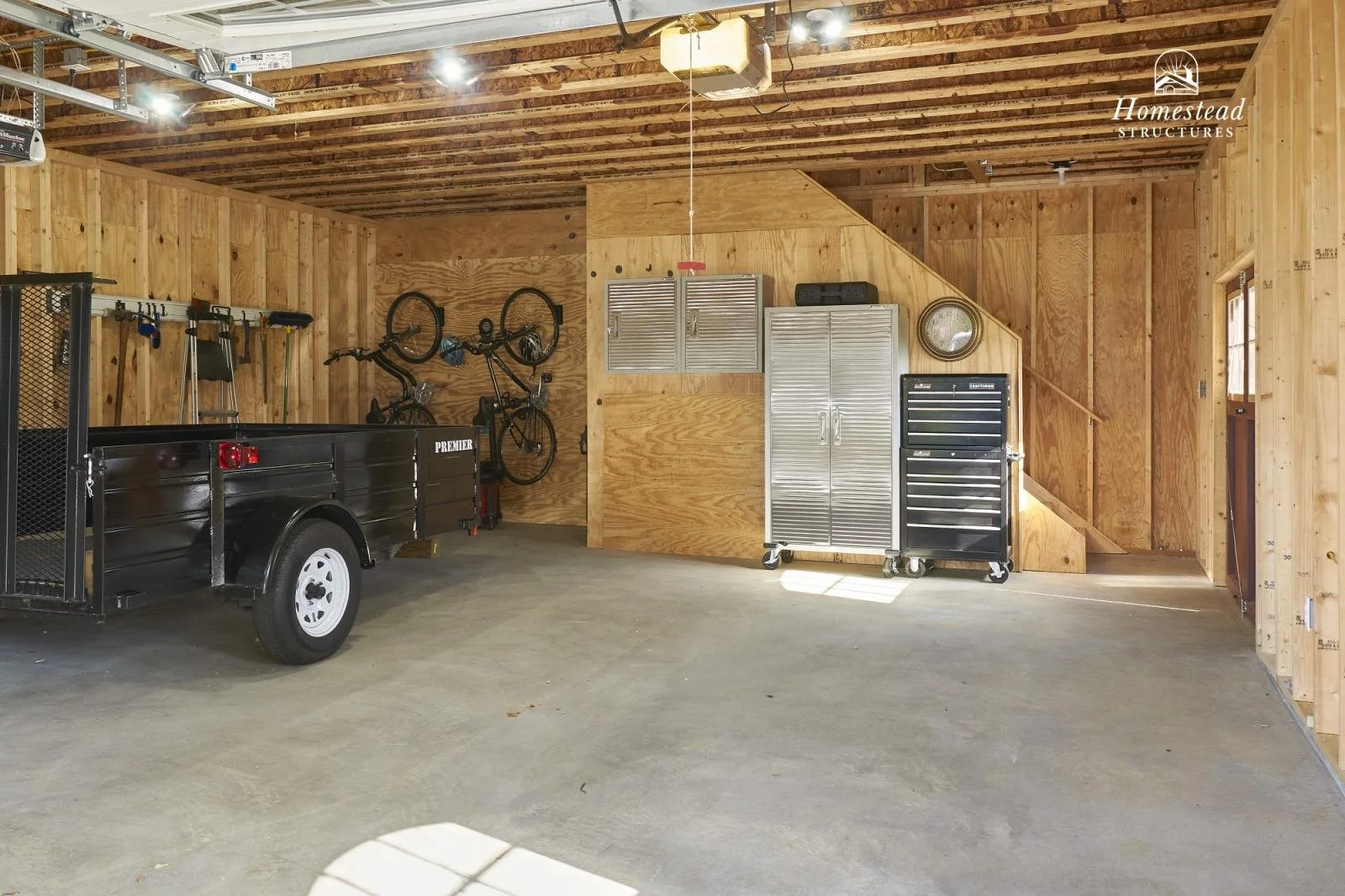 Clean, organized garage with wooden walls and ceiling, featuring a black utility trailer, bicycles mounted on the wall, gray metal cabinets, a clock, and a black tool chest.