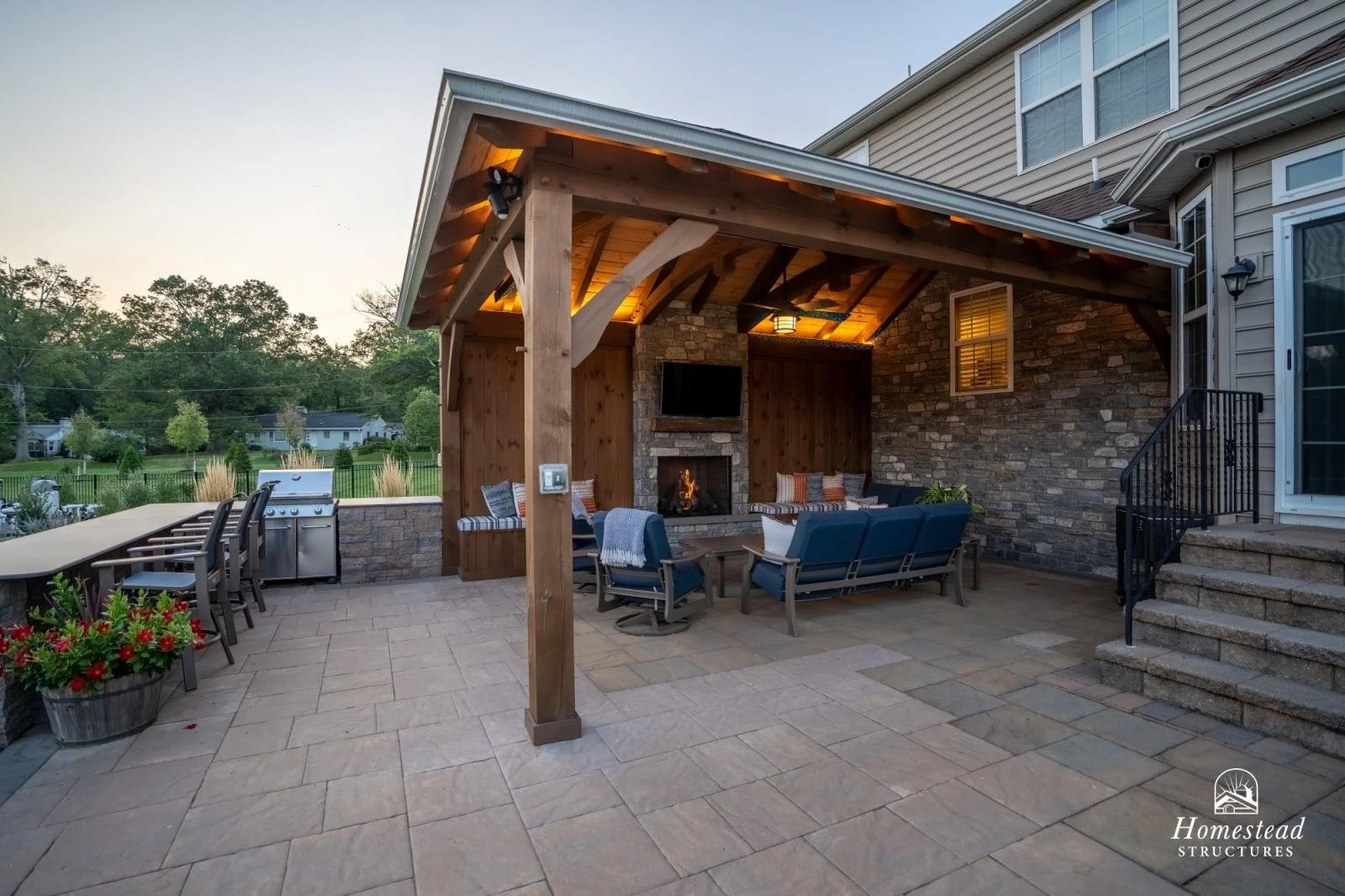 Backyard patio with a covered outdoor seating area, fireplace, flat-screen TV, and outdoor kitchen, surrounded by a stone wall.