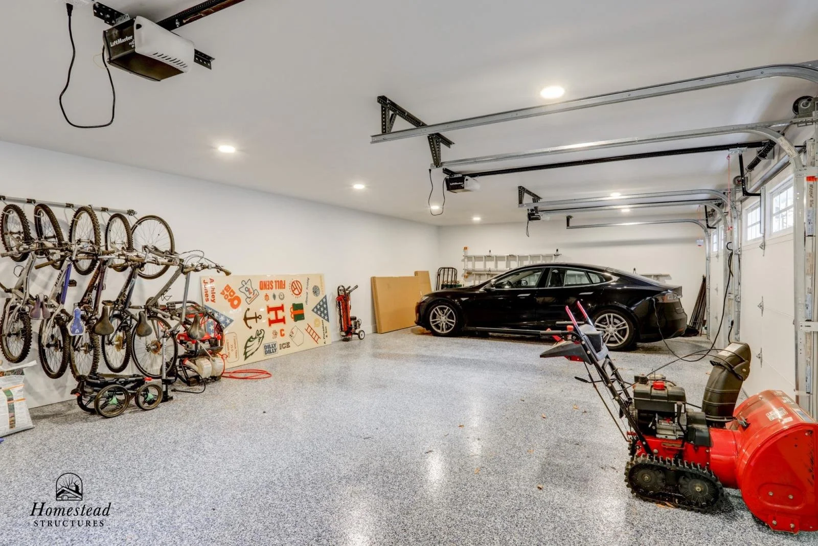 A spacious garage with a black car parked inside, bicycles mounted on the left wall, a snow blower in the foreground, and various tools and boards along the back wall. The garage has a clean, organized appearance with overhead lighting.