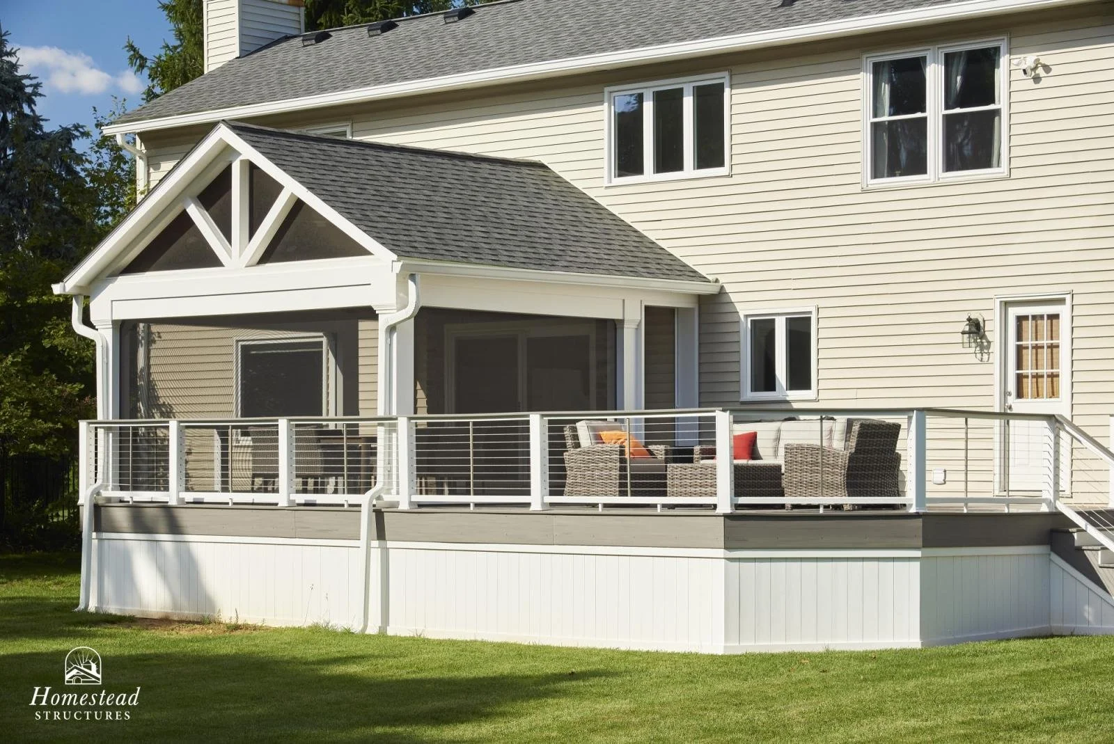 A house with a large white deck featuring outdoor furniture, including wicker chairs with cushions, attached to beige siding. The house has multiple windows, a small door leading onto the porch, and a gable roofed entryway. A well-maintained green la