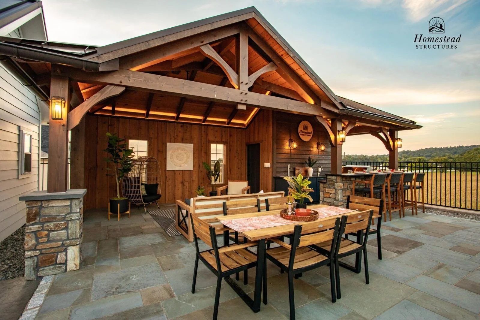 Outdoor patio with wooden furniture, a dining table with four chairs, a seating area with a sofa and a lounge chair, a built-in bar area with barstools, and string lights, with a view of a field and trees in the distance.