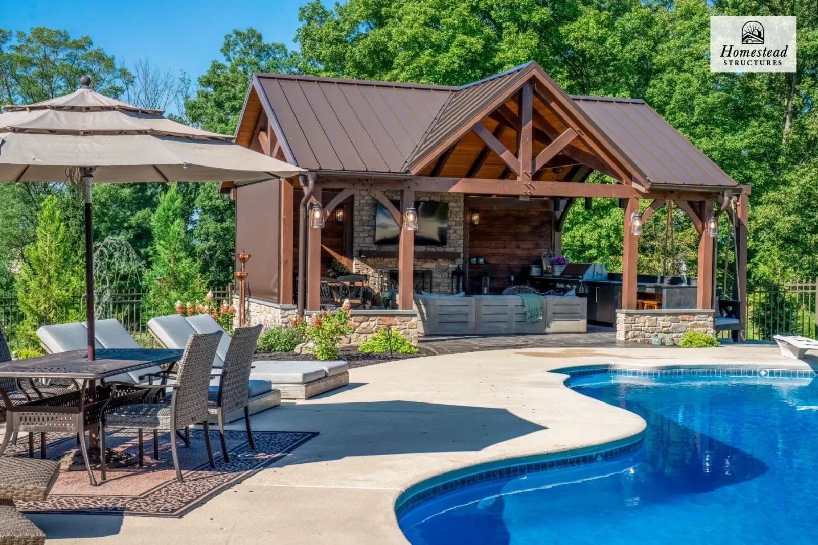 Outdoor backyard with a swimming pool, patio furniture, umbrella, and a wooden pavilion with a TV and outdoor kitchen, surrounded by trees.