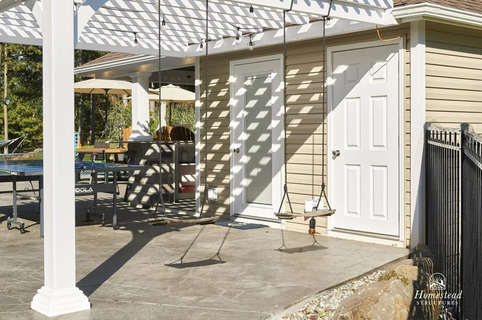 Backyard patio with two white doors, a swing, table tennis table, and a shaded pergola with string lights.
