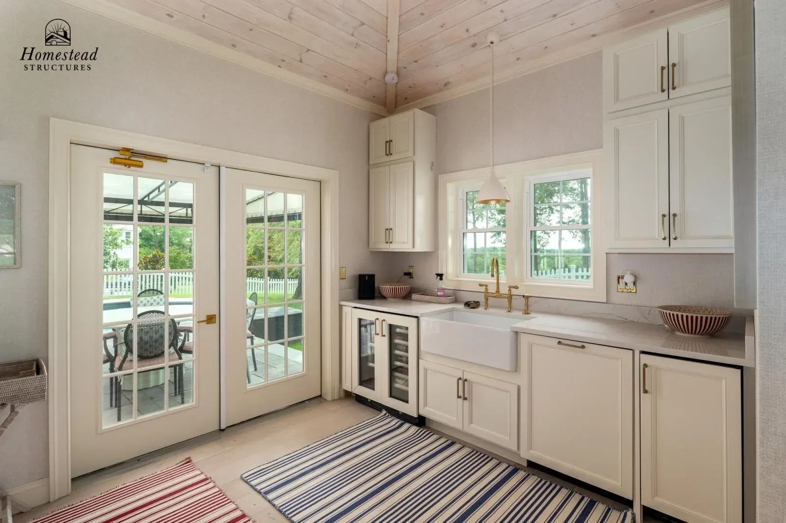 Kitchen with white cabinets, a farmhouse sink, gold hardware, a window, and glass French doors leading to an outdoor patio with a dining table and chairs.