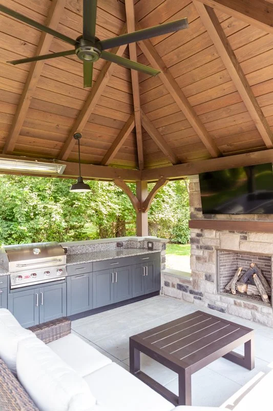 Outdoor pavilion with a wooden ceiling, ceiling fan, and mounted TV, featuring a built-in grill, a gray cabinet with a countertop, and a patio with seating and a coffee table, surrounded by trees.