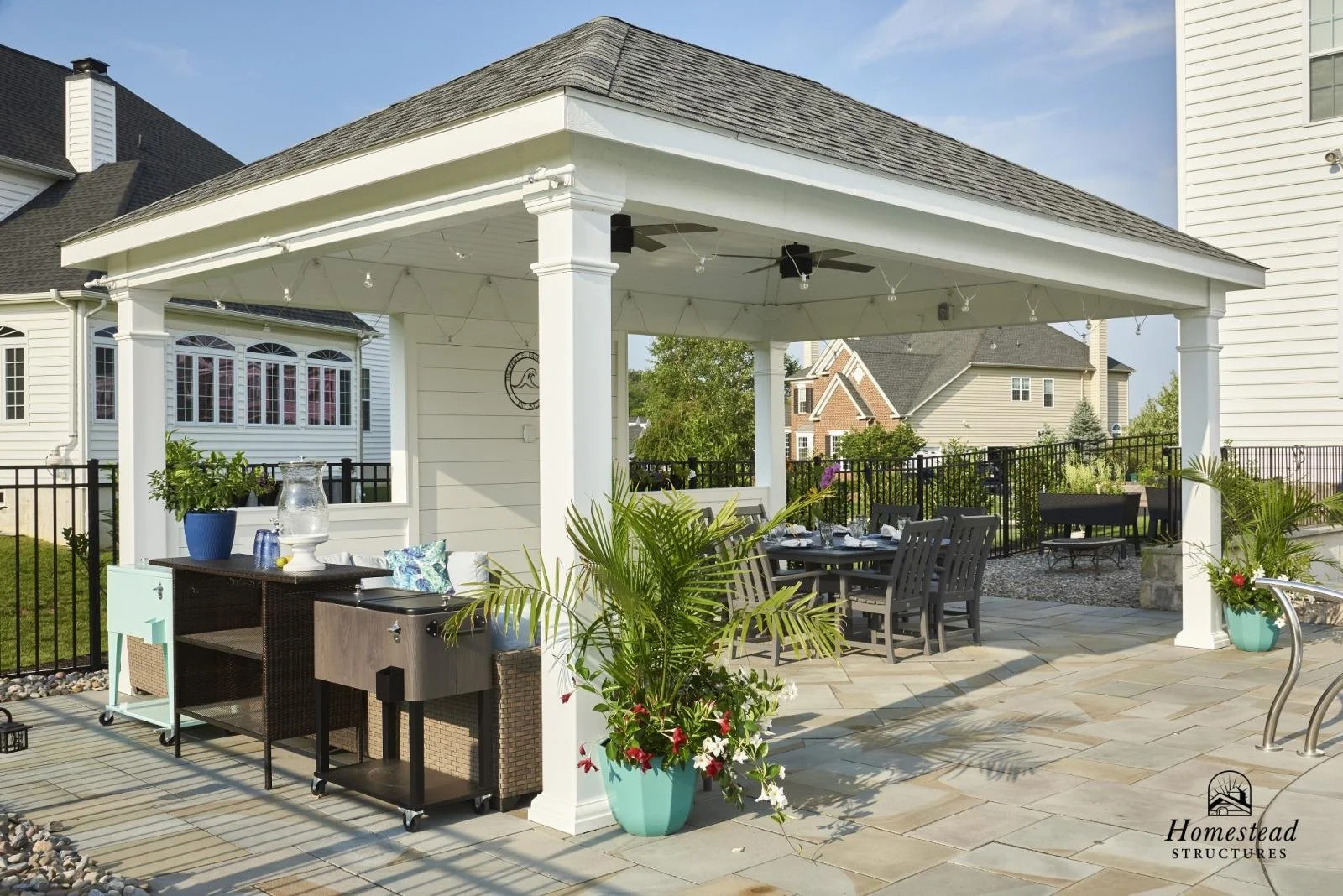 Outdoor patio area under a white gazebo with a dining table, chairs, potted plants, and outdoor furniture, with neighboring houses and a black metal fence in the background.