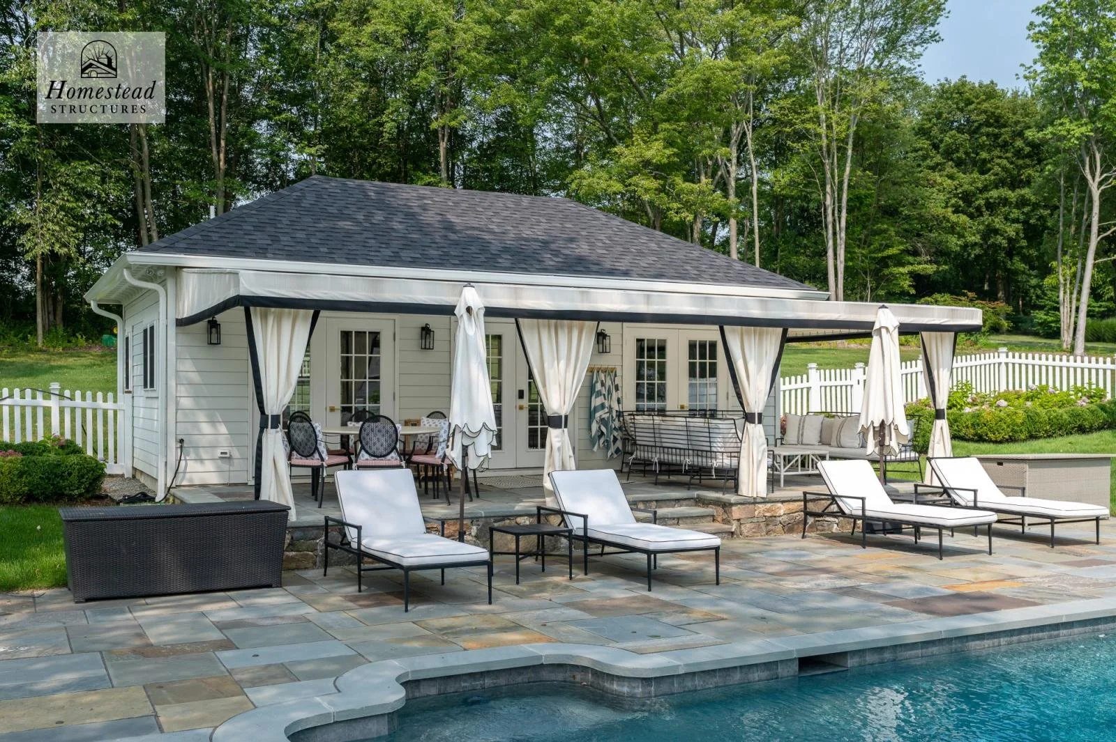 Backyard with a pool and a pool house, outdoor seating, patio furniture, and chairs with umbrellas, surrounded by a white fence, trees, and greenery.