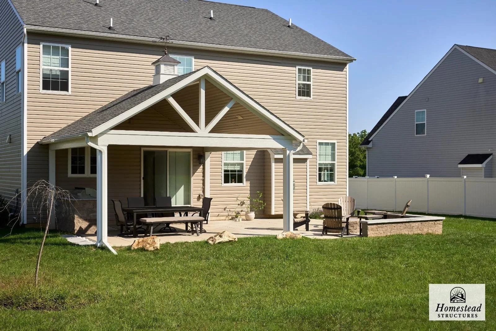 Backyard with a covered patio with outdoor furniture, a small fire pit, a white fence, and a two-story beige house with multiple windows.