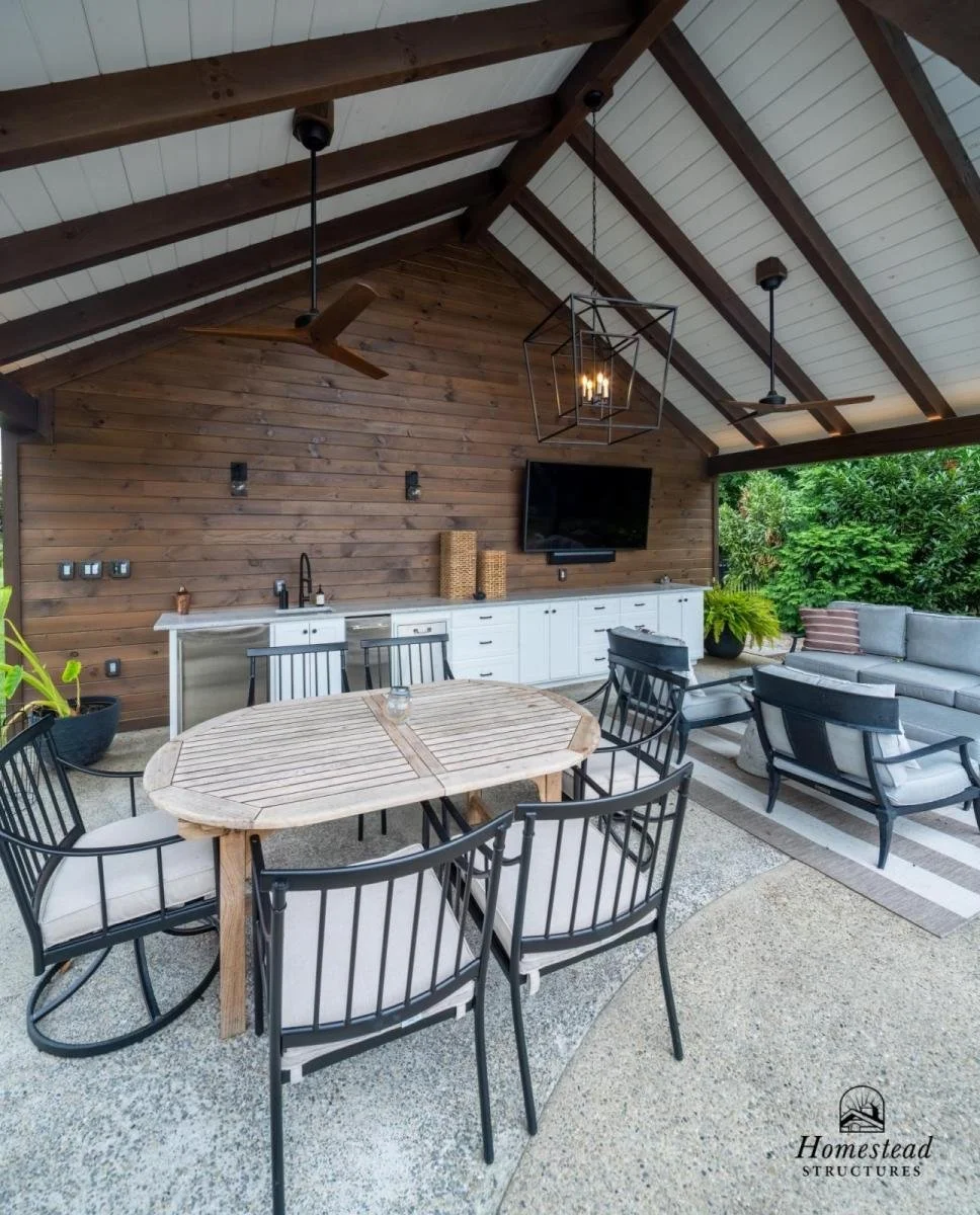 Outdoor covered patio with wooden ceiling and wall, black ceiling fans, a flat-screen TV, white cabinets, a round wooden dining table with six black chairs, a gray outdoor sofa with pillows, and lush greenery in the background.