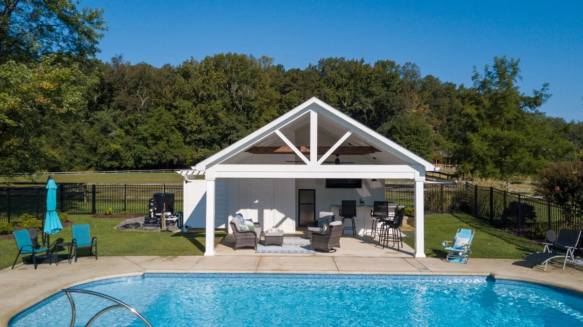 Backyard with a swimming pool, poolside chairs, and a covered patio area with outdoor furniture, barbecue grill, and barstools, surrounded by a black metal fence and trees in the background.