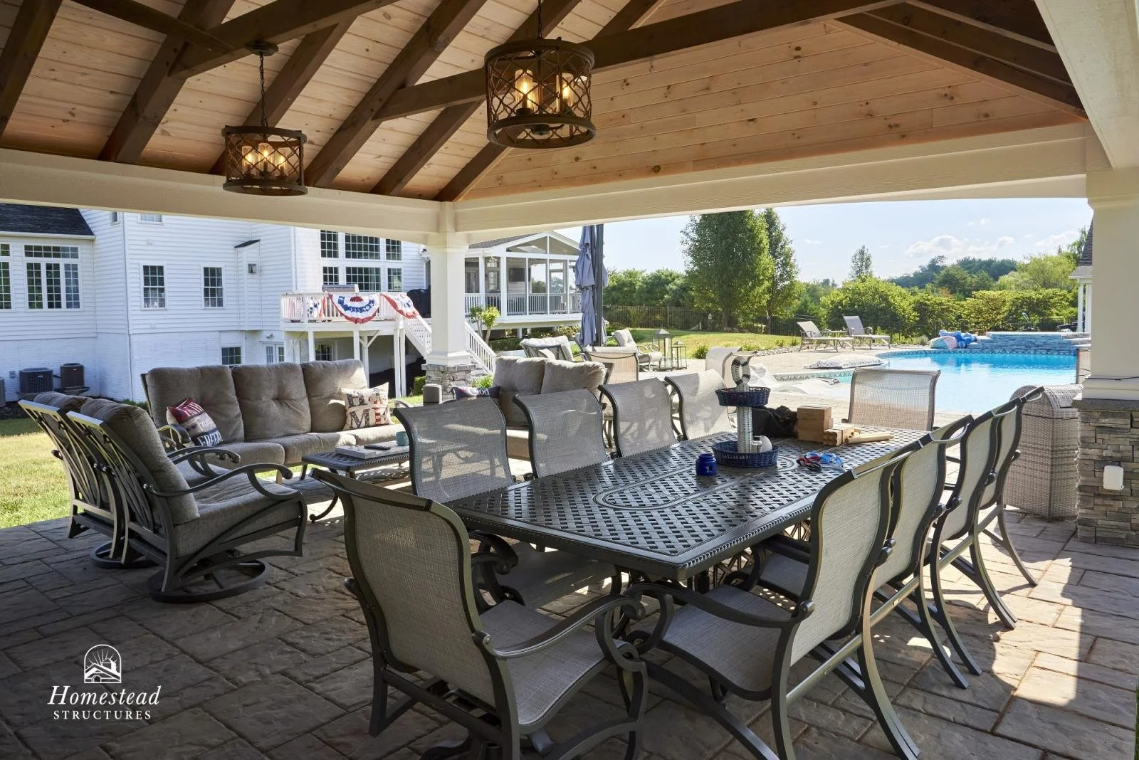 Covered outdoor patio with a dining table and chairs, a seating area with cushioned chairs, overlooking a backyard with a swimming pool, lounge chairs, and lush green trees.