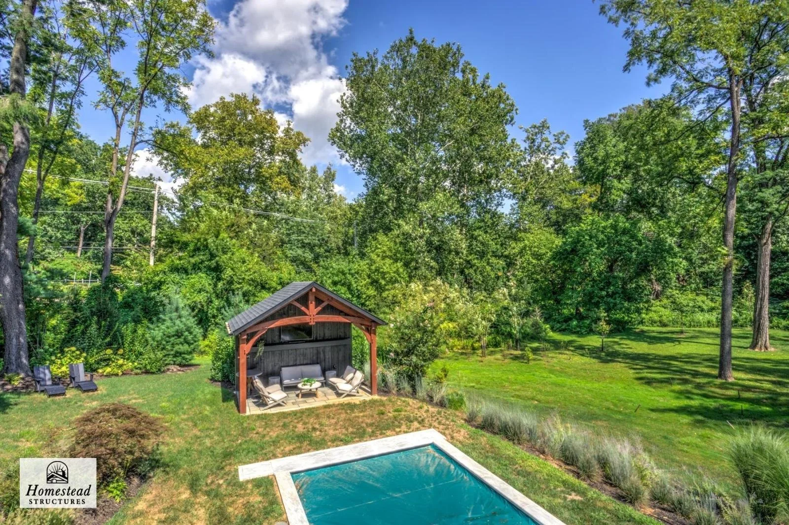 Backyard with a small covered seating area, a swimming pool, lush green grass, and tall trees under a partly cloudy sky.