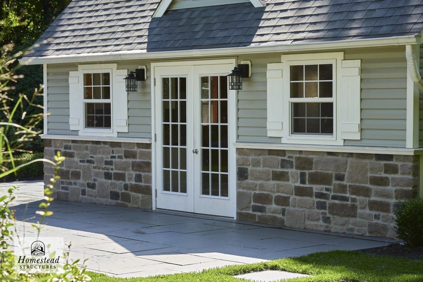 A small house with a stone foundation, beige siding, and two white-framed windows with shutters. There are two black outdoor wall lights beside a set of French doors. A concrete patio is in front, and there is greenery on the sides.
